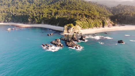 La-Luz-Del-Sol-De-La-Tarde-Sobre-Las-Hermosas-Aguas-Prístinas-De-La-Playa-De-Monro-En-La-Isla-Sur,-Nueva-Zelanda-Con-Un-Exuberante-Paraíso-Tropical-En-El-Fondo---Drone-Aéreo