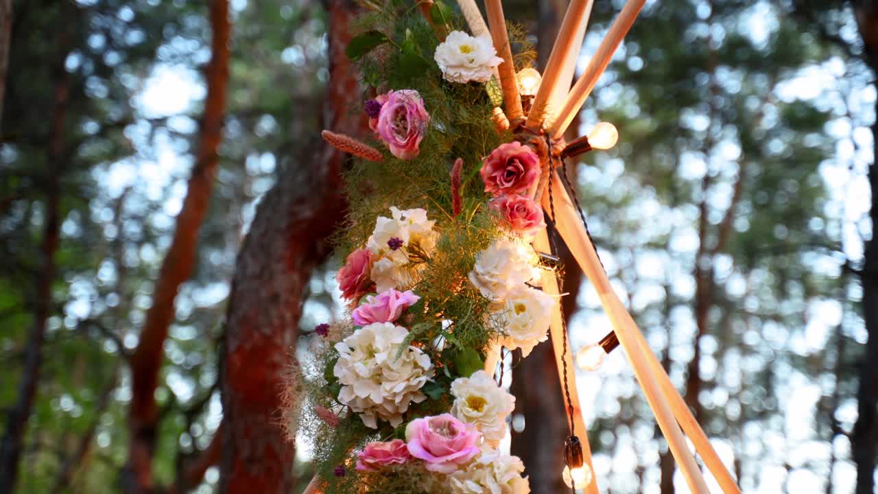 Bohemian tipi wooden arch decorated with pink roses, candles on carpet, pampass grass, wrapped in fairy lights on outdoor wedding ceremony venue in pine forest at night. Floristic compositions