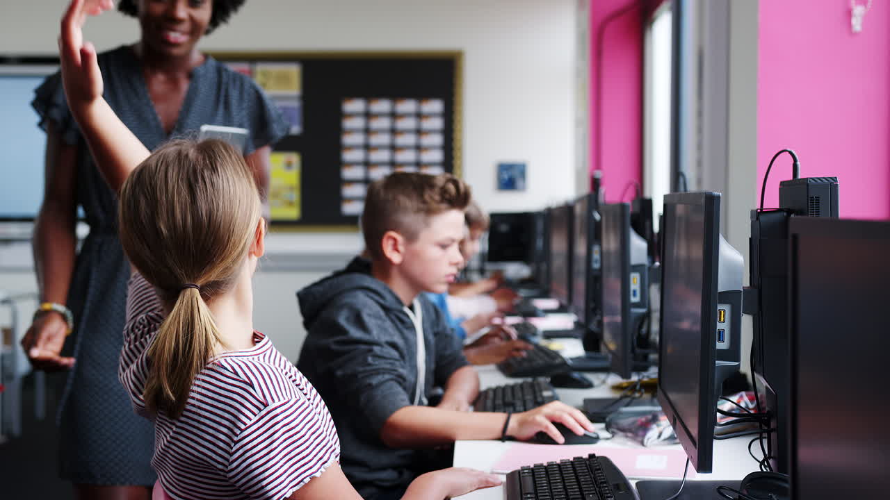 Teacher Helping Female Pupil In Line Of High School Students Working at Screens In Computer Class