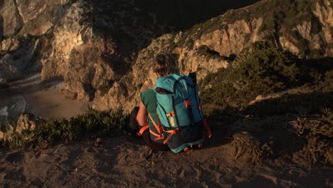 back view of female tourist with backpack sitting at cliff