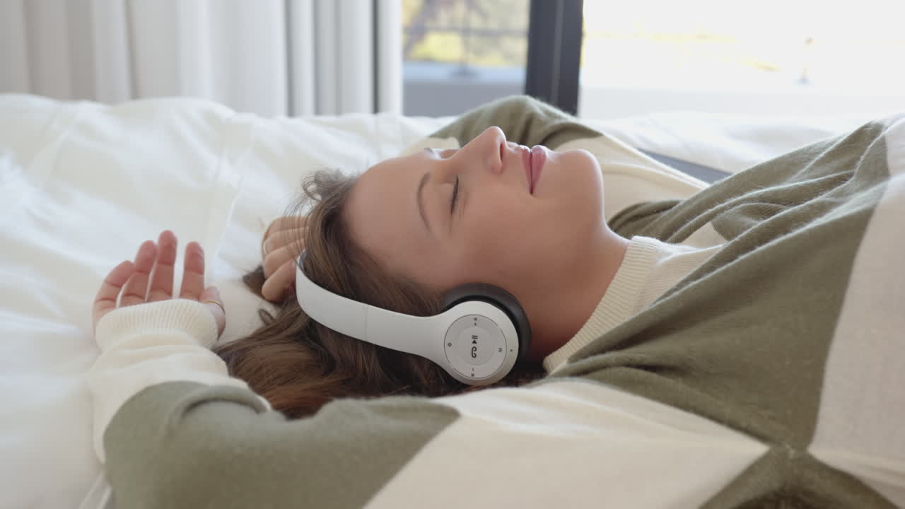 Relaxing on bed, young woman listening to music with headphones, smiling peacefully