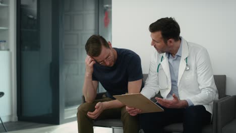 Caucasian-male-doctor-and-patient-sitting-discussing-documents-in-corridor-of-health-center