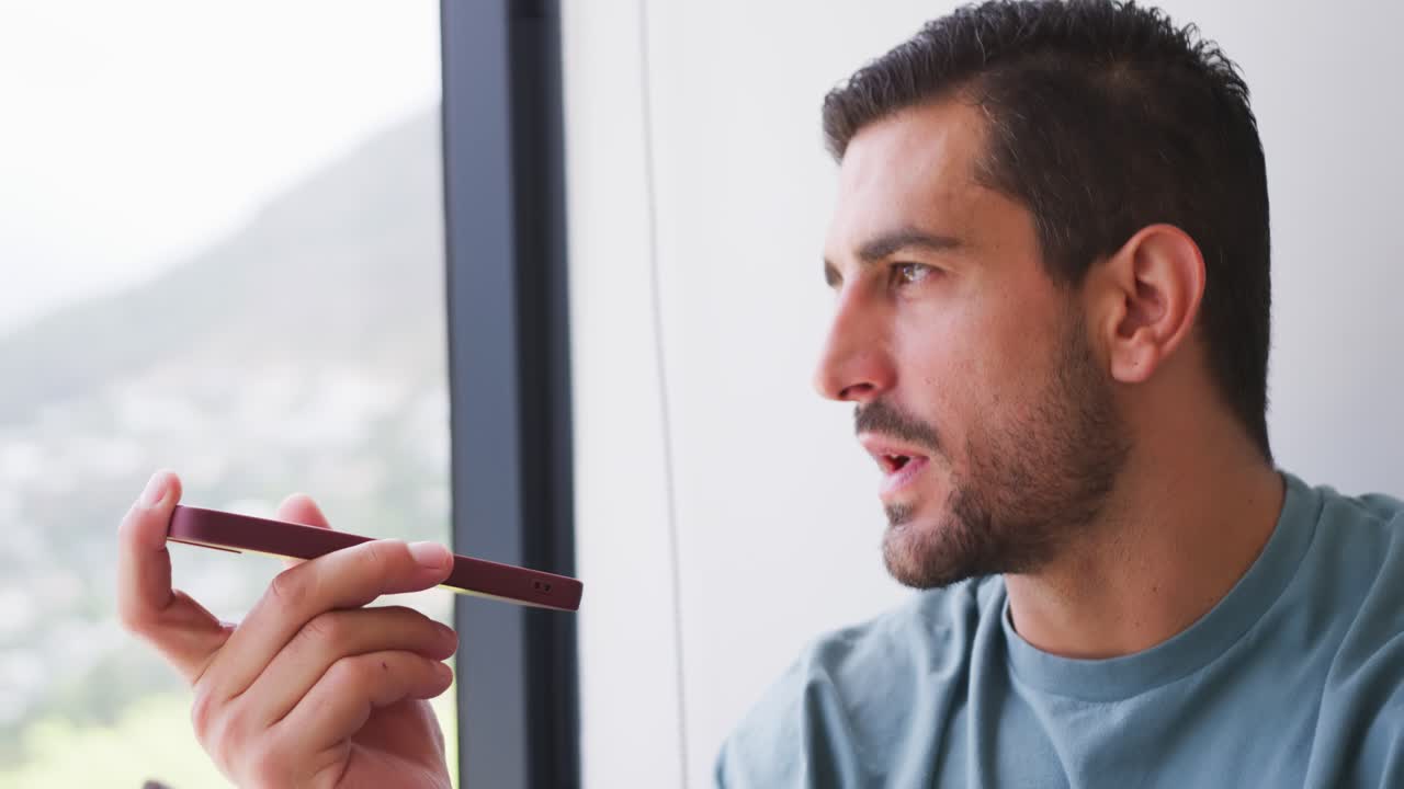 Uomo adulto seduto vicino alla finestra che alza lo smartphone rosso per parlare, mentre parla, ascolta e guarda fuori.