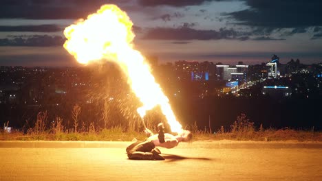 Young blond male kneels with his left side to the camera reclines as he breathes fire