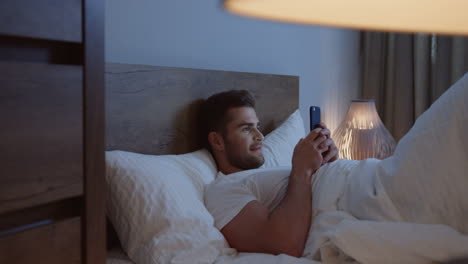 hombre caucásico acostado en la cama, charlando en el teléfono inteligente a altas horas de la noche antes de dormir