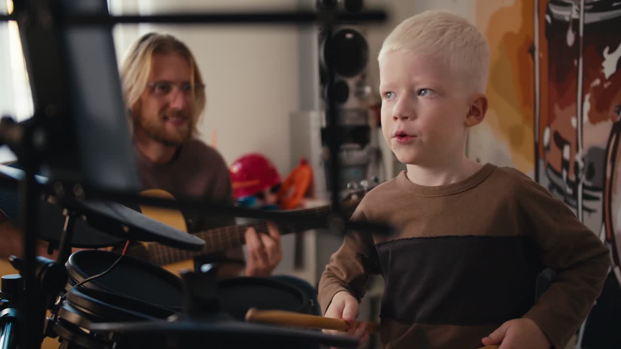 하 머리와 파란 눈을 가진 행복한 알비노 소년은 특별한 막대기를 사용하여 전기 음악 세트에서 연주하고, 수염과 안경을 쓴 금발 남자가 아들을 도와 기타를 함께 연주합니다.