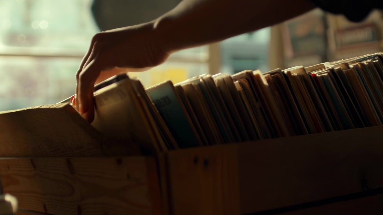 Una mano illuminata caldamente si libra sopra una scatola di stoccaggio affollata piena di dischi in vinile in verticale, creando una scena interna nostalgica con ombre morbide e una messa a fuoco ridotta.