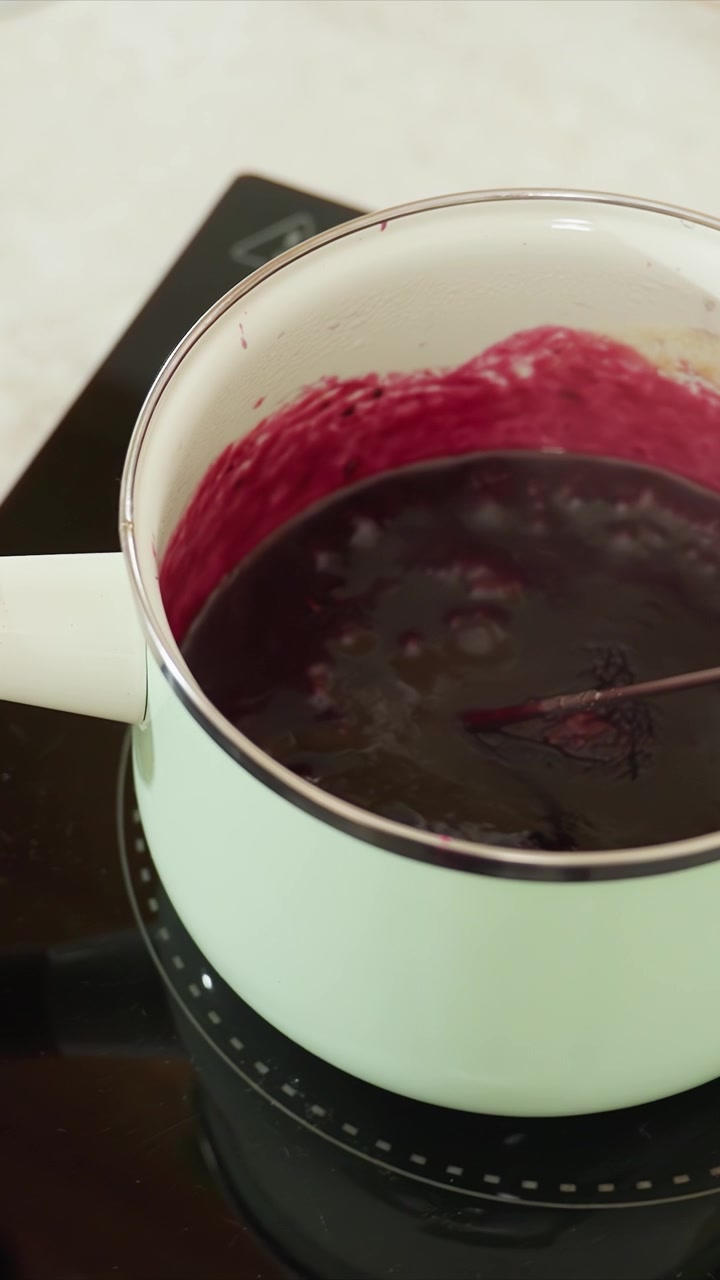 Primo piano di una pentola verde chiaro con manico bianco su piano cottura a induzione. Un cucchiaio di legno mescola il composto mostrando un sobbollire continuo. Il piano di lavoro della cucina è visibile sullo sfondo.