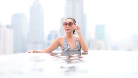 sensual slim asian thai woman with vintage sunglasses relaxing in luxurious infinity pool with incredible view of bangkok cityscape in background