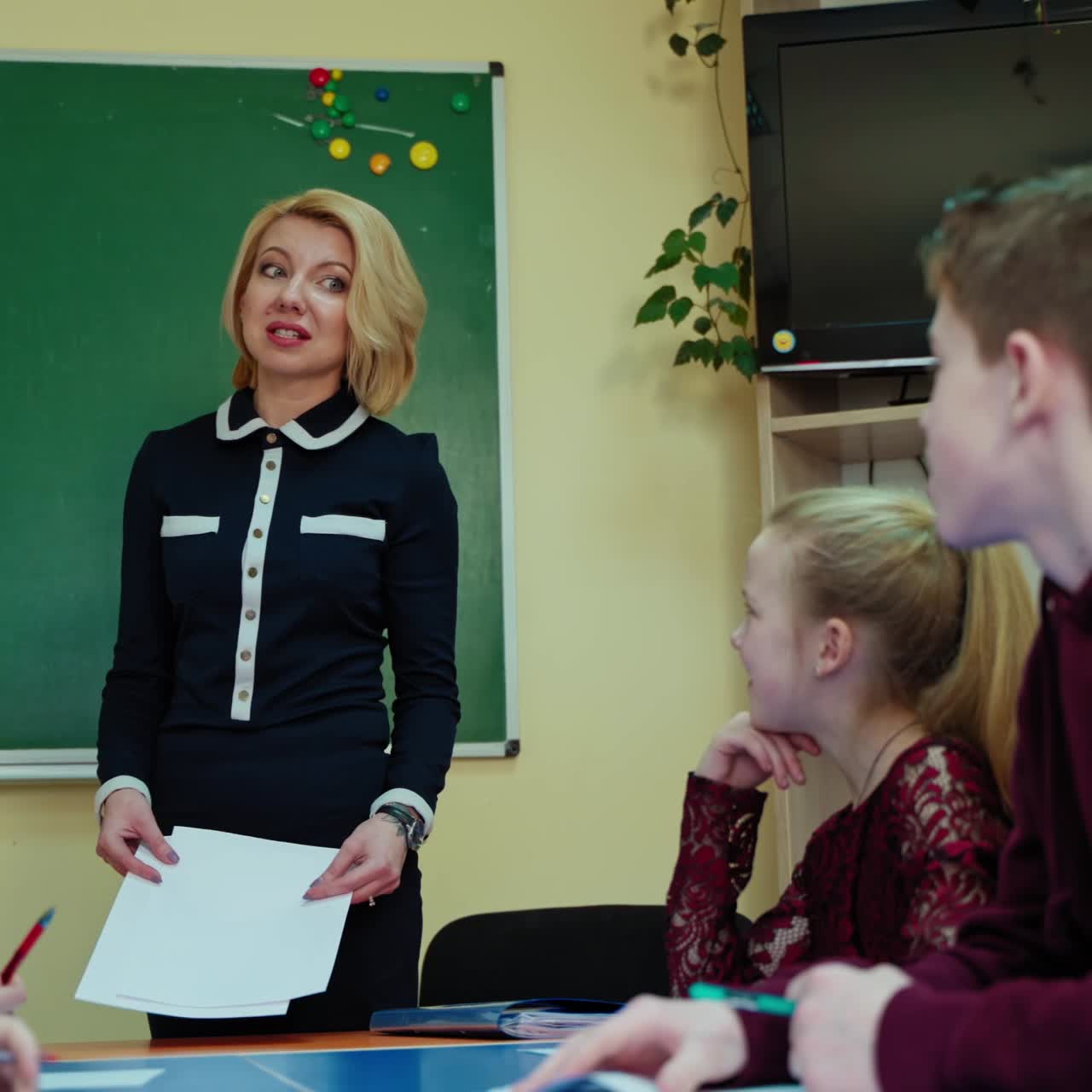 Back to school. Beautiful teacher talking with school children on the background of blackboard. Student asking questions to the teacher in the classroom.