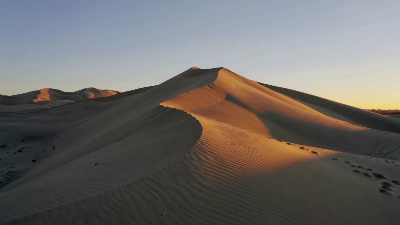 Drone in movimento che rivela una duna a mezzaluna illuminata dal sole al tramonto nel deserto, mostrando increspature e impronte.
