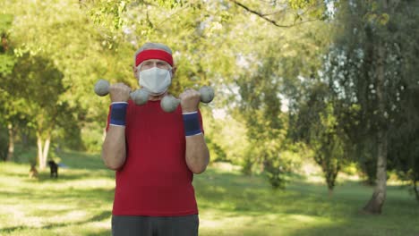 senior man making fitness exercises with dumbbells during coronavirus virus pandemic alone in park