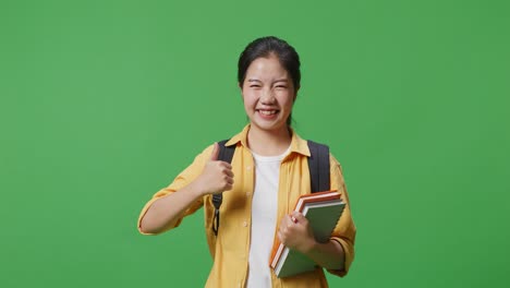 녹색 스크린 배경 스튜디오에 서 있는 동안 웃고 엄지손가락을 보여 카메라에 제스처를 하는 배과 몇 권의 책을 가진 아시아 여성 학생