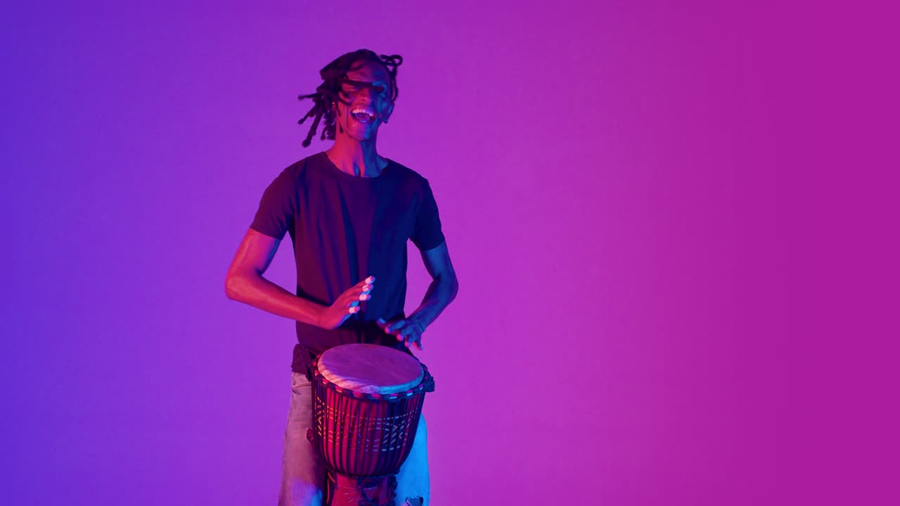hombre negro se ríe tocando un tambor africano y bailando en el estudio. el músico bate el ritmo en el tambor con las manos y baila alegremente en un fondo de gradiente