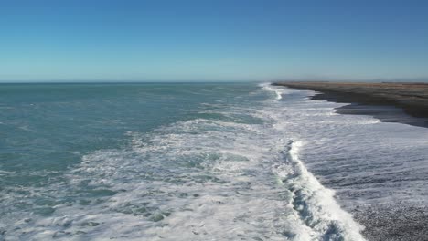 Low, slow aerial above white-water of incoming waves from beautiful turquoise ocean on a clear, sunny day - Birdlings Flat, Beach