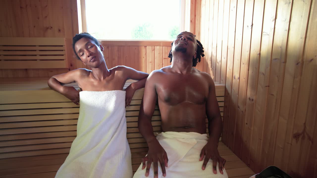 Coppia afroamericana seduta su una panca sotto un pannello, il calore della sauna li fa appoggiare con gli asciugamani, vicino alla stufa.