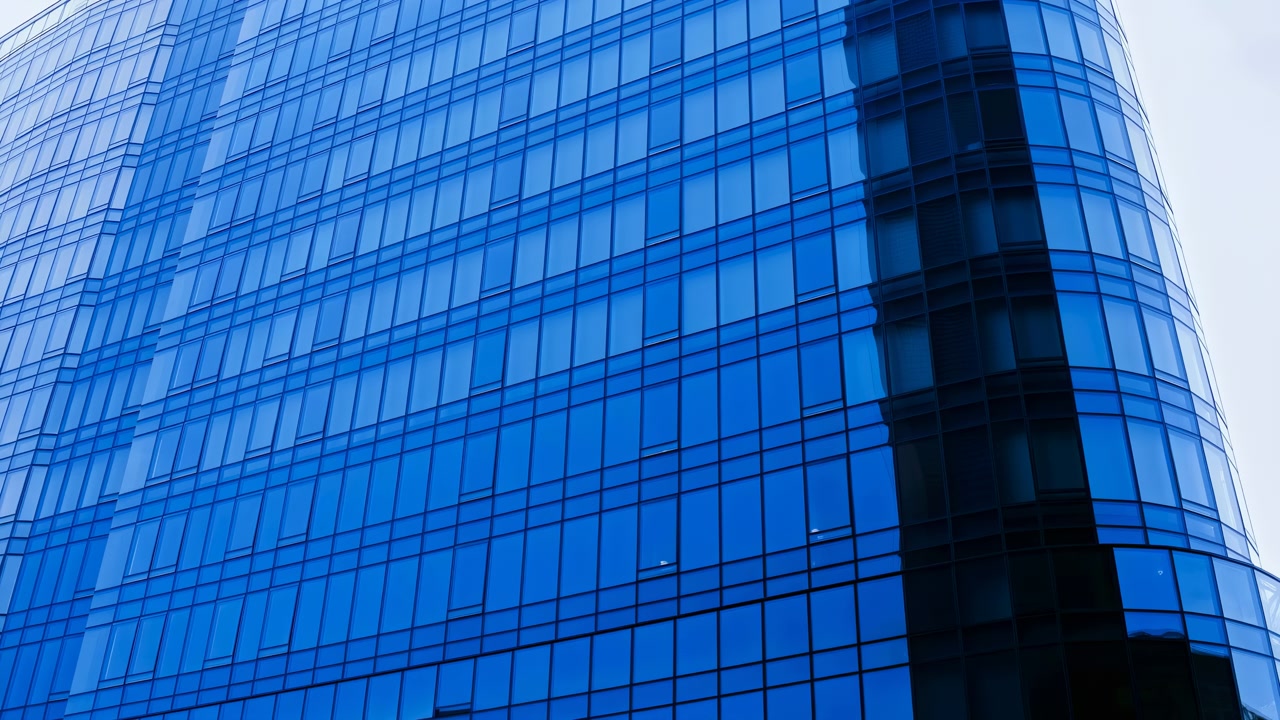Vista ravvicinata obliqua verso l'alto della facciata di un grattacielo per uffici con finestre ripetute in vetro blu, angolo curvo scuro e riflessi cangianti che rivelano un'elegante architettura aziendale in movimento