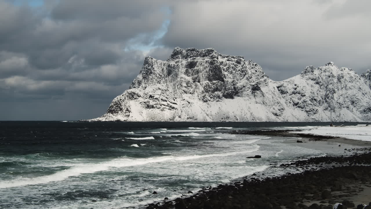 Sjøbølger som skvulper mot land med snødekte, røffe fjell og overskyet vær i bakgrunnen