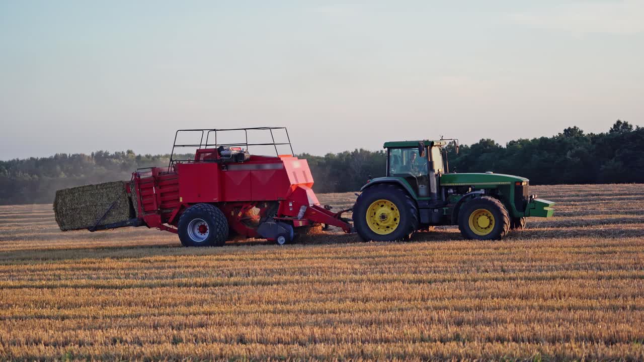 Harvesting dry grass. Agricultural works in the field. Tractor is pressing hay into square bale on the natural yellow field background.
