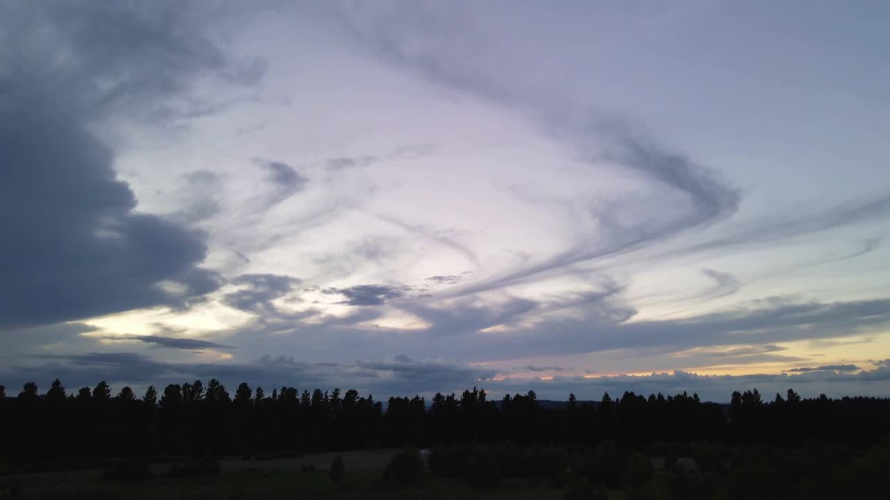 Una massa nuvolosa vorticante accentua bande a spirale sopra gli alberi al crepuscolo, con il sole che si avvicina all'orizzonte e causa un bagliore.