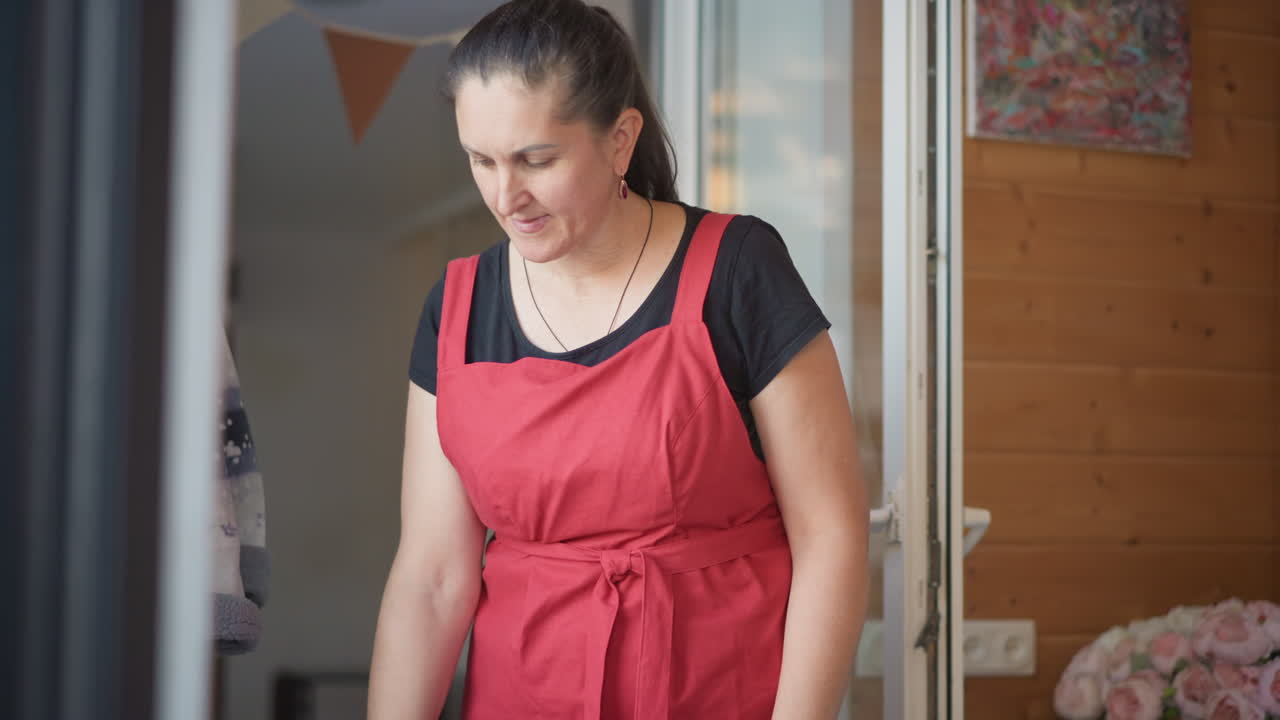 Mulher cozinheira entrando pela porta com avental vermelho, pisando em uma cozinha de madeira aconchegante com bandeirolas e uma planta, inclinando-se para pegar objetos perto da soleira, com luz natural e uma rotina doméstica tranquila.