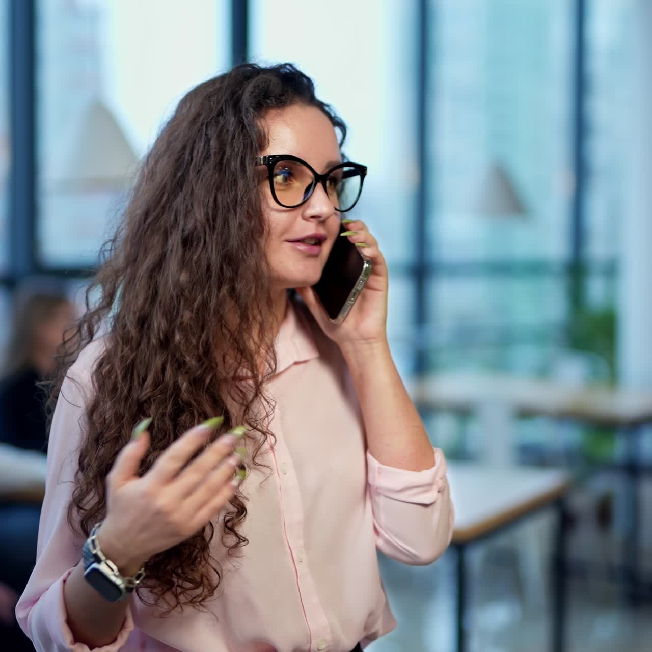 Attractive lady making a phone call standing in office. Dark-haired woman talking and waving with her hand. Close up portrait