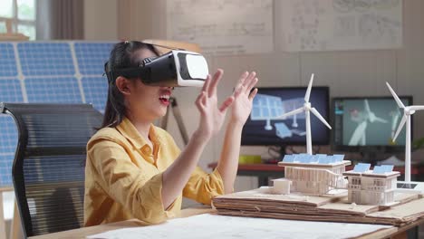 side view of smiling asian woman working with a model of small house roof solar panel wearing vr and showing touching gesture