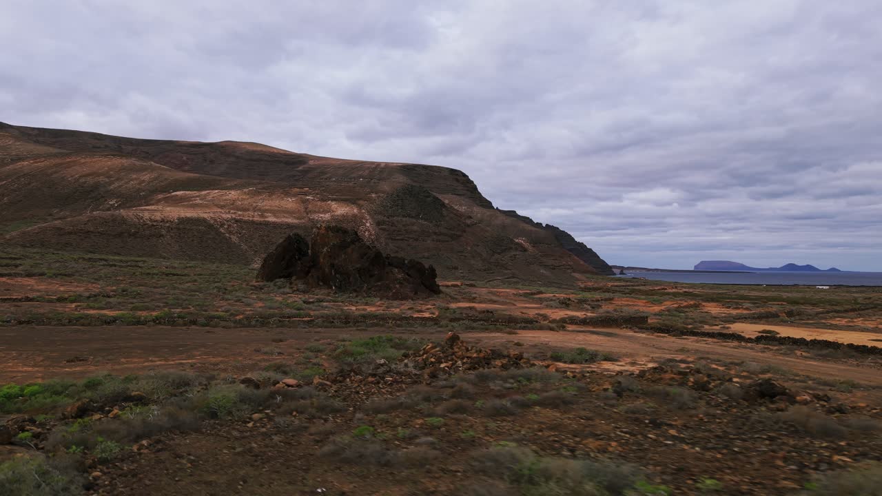 Paesaggio vulcanico arido a Lanzarote, caratterizzato da una pianura, montagne e vegetazione sparsa sotto un cielo nuvoloso.