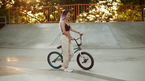 Attractive-stylish-girl-with-dreadlocks-walks-with-bmx-bike-in-skatepark-in-a-recreation-park.-Young-stylish-woman-riding-a-bike