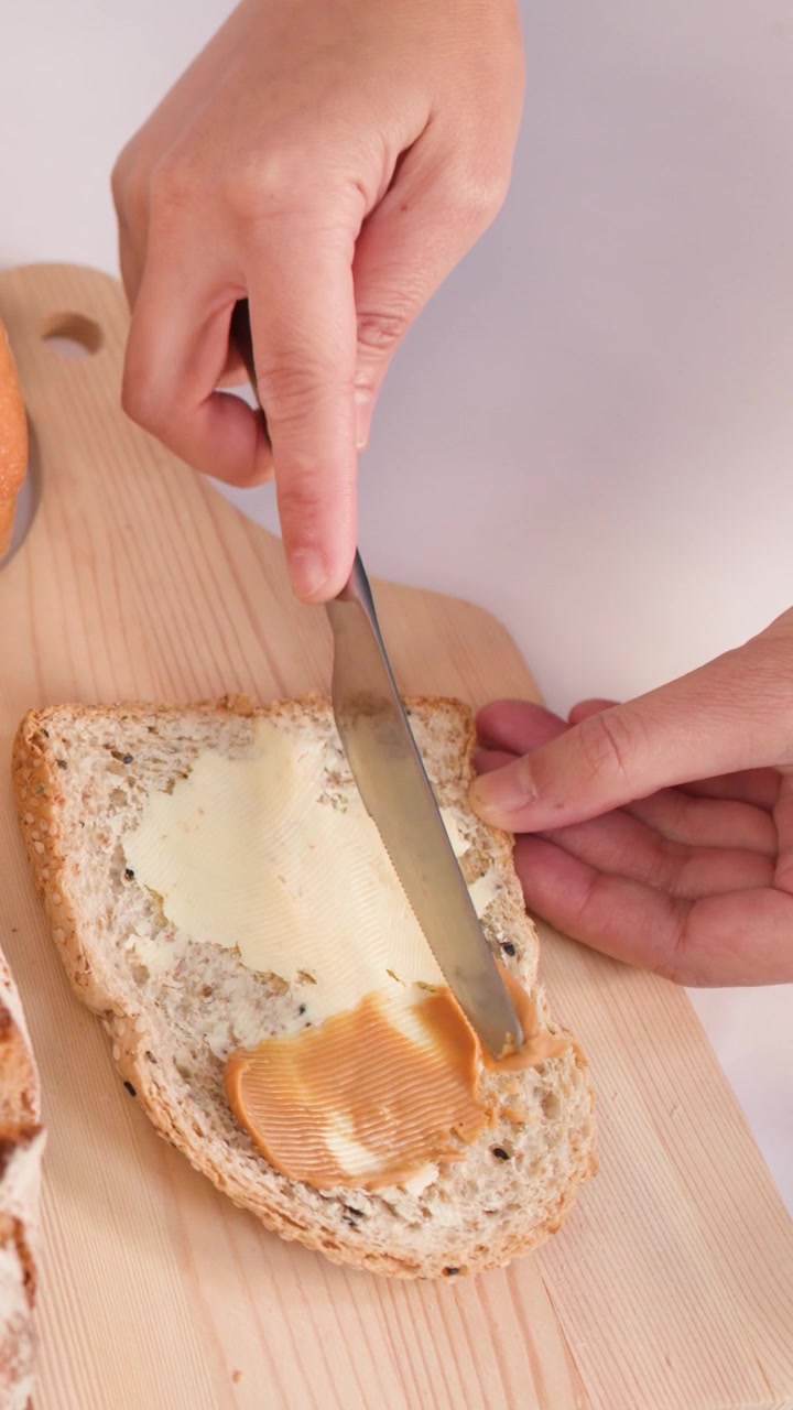 Hands spread peanut butter over buttered multigrain bread on a wooden board with assorted loaves.
