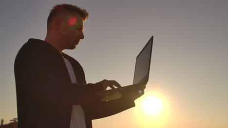 middle plan: a male freelancer typing on a laptop keyboard at sunset overlooking the city while standing on the roof of a skyscraper. the businessman works remotely and controls the work of the company