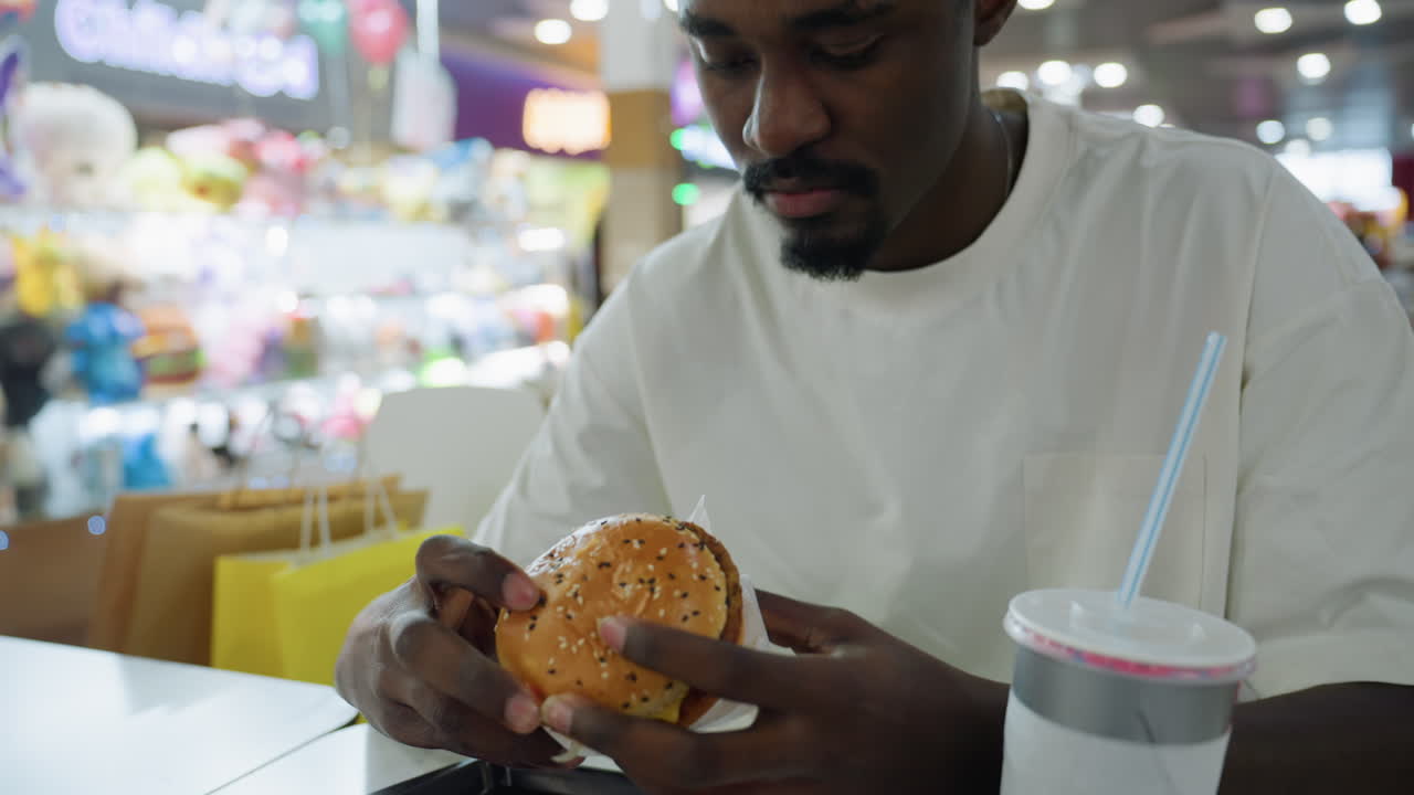 Young man wearing white shirt sits in indoor food court holding burger with both hands ready to take bite while cold drink in disposable cup with straw rests on tray near packaging and paper napkin