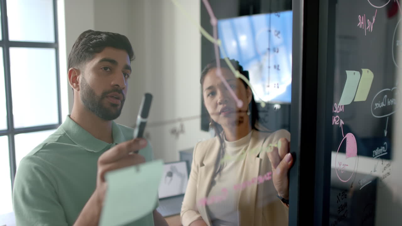 Discussing project strategy, colleagues writing on glass board in modern office