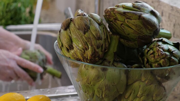 Woman washing artichokes in stainless steel sink with metal faucet. Cooking artichokes at kitchen