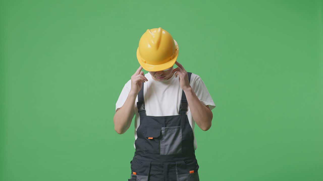 un trabajador asiático con gafas de protección y casco de seguridad tiene dolor de cabeza mientras está de pie en el estudio de fondo de la pantalla verde