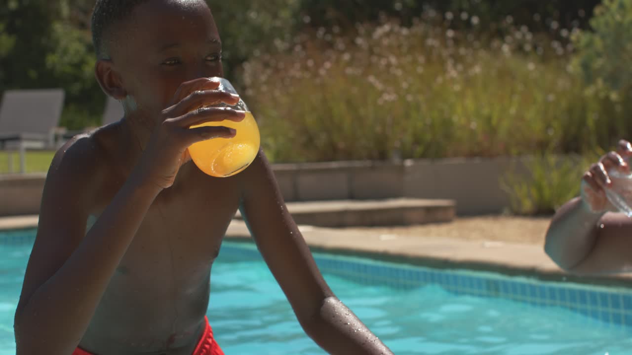 African American father and teen son shirtless clinking glasses poolside, son sipping orange drink