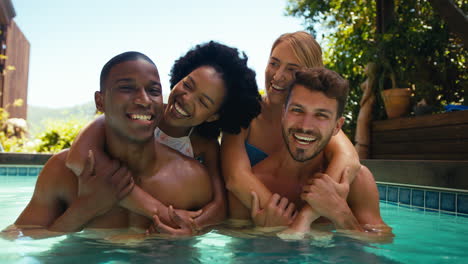 portrait of multi-cultural friends on holiday in swimming pool with men giving women piggybacks