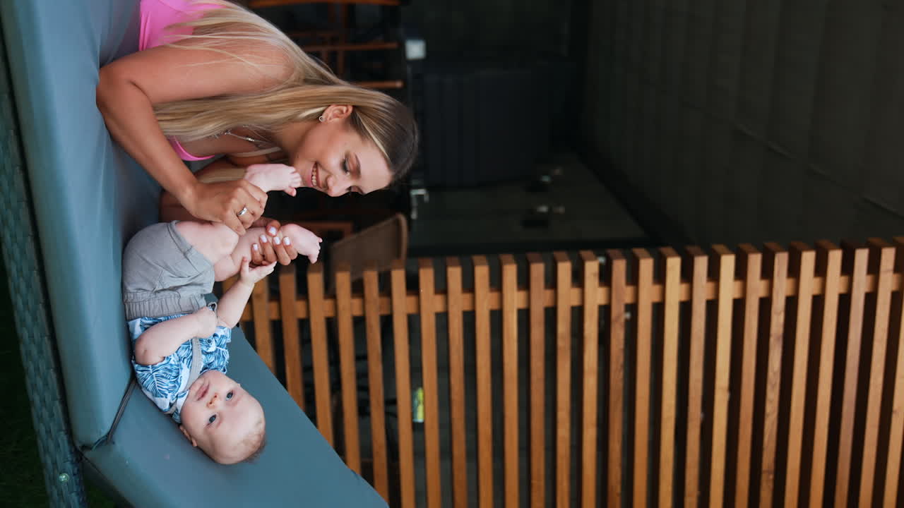 Beautiful blonde woman playing with her baby lying on the deck chair. Mom is waving kid's little feet, tickling and kissing them. Vertical screen.