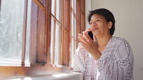 Mujer-Mayor-Afroamericana-Sonriendo-Mientras-Habla-Por-Teléfono-Inteligente-Cerca-De-La-Ventana-En-Casa