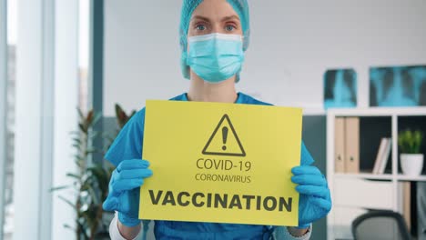 close up of beautiful female specialist doctor in protective gloves and medical mask holding in hands yellow paper card showing notification about covid 19 vaccination, coronavirus pandemic