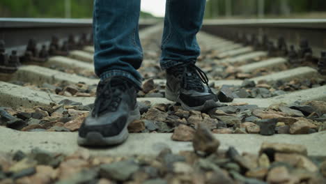 close leg view of someone wearing jeans trousers and canvas shoes, walking on a railway with stones, the background includes a blur of trees and electric poles on either side