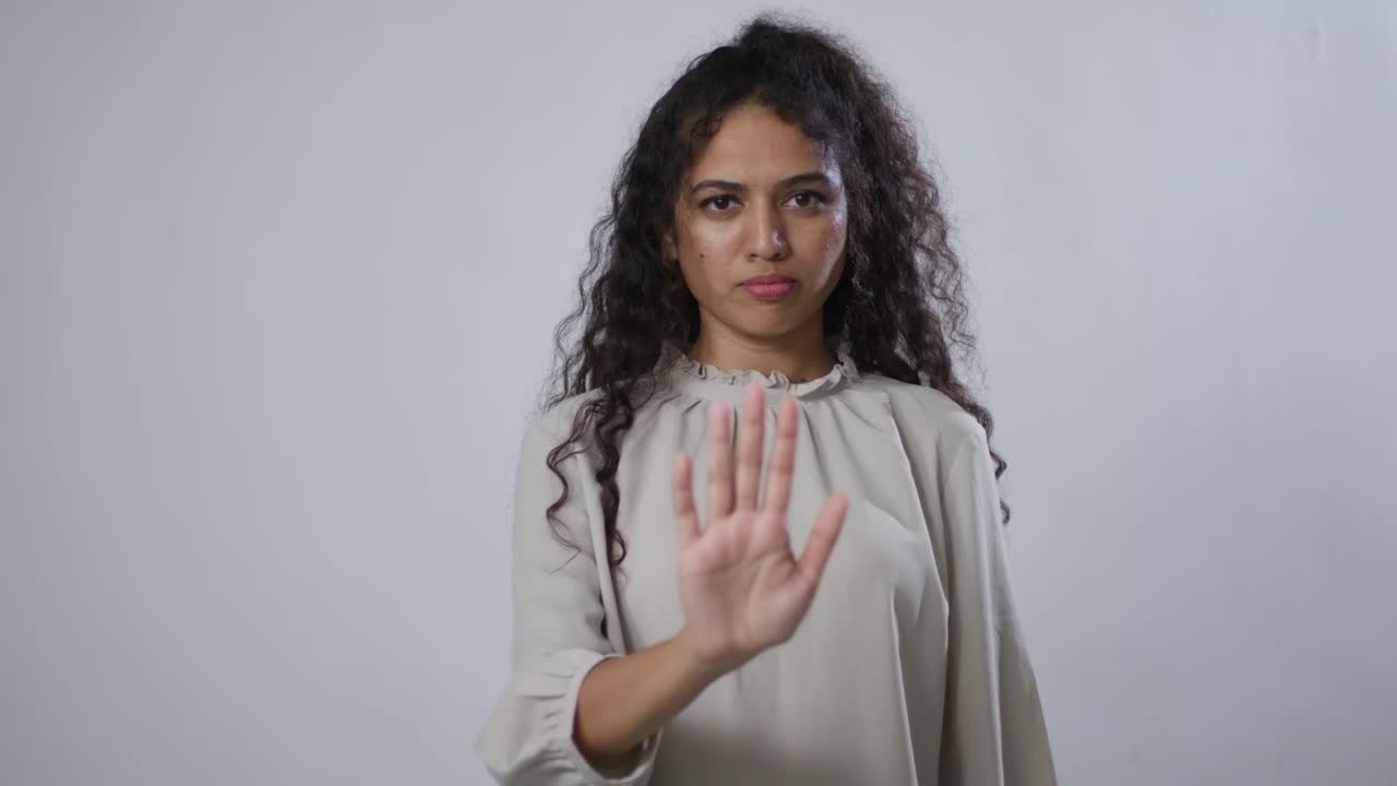 A South Asian woman with curly hair points to the viewers and holds up her hand with her palm facing out, as if to say, "Stop"