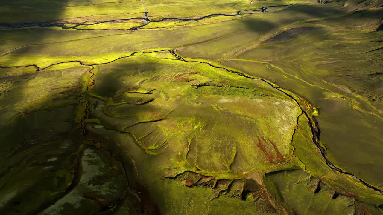 Aerial sweep over vivid Icelandic moss plains and fractured volcanic ridges, revealing organic patterns shaped by erosion and ancient lava flows under soft afternoon light