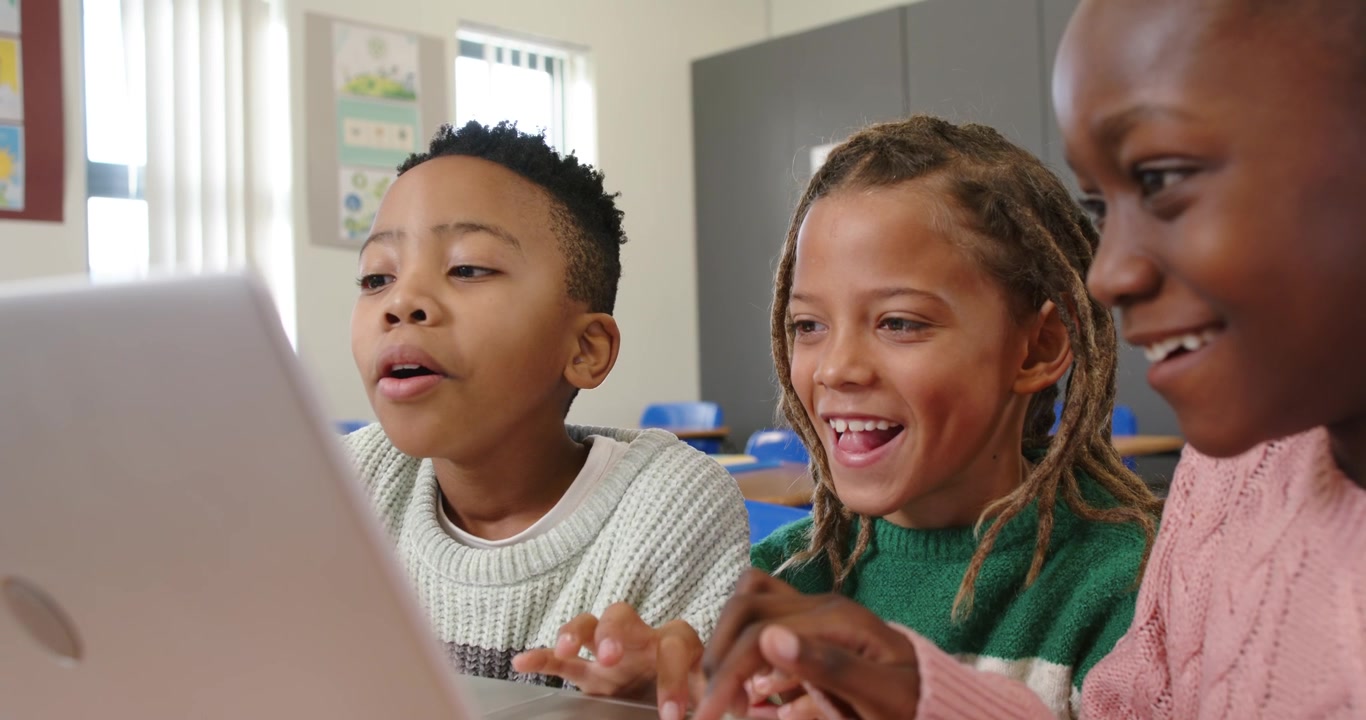 Diverse children leaning over laptop at school desk, reacting to on-screen task, typing classwork