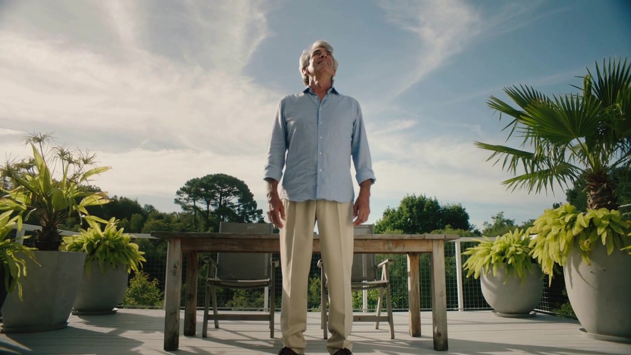 Un uomo anziano si inclina, indossando una camicia blu e pantaloni beige, guardando il cielo soleggiato su una terrazza con fioriere.