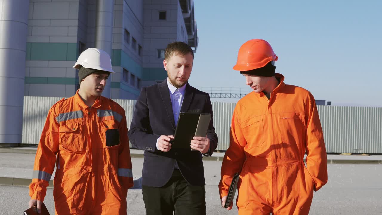 ingeniero en traje y dos trabajadores en uniforme naranja y cascos están caminando a través de la instalación de construcción. filmado en 4k