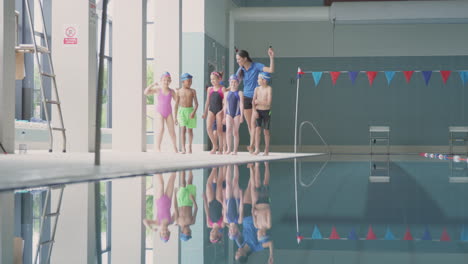 Female-Coach-Walks-Children-In-Swimming-Class-Along-Edge-Of-Indoor-Pool