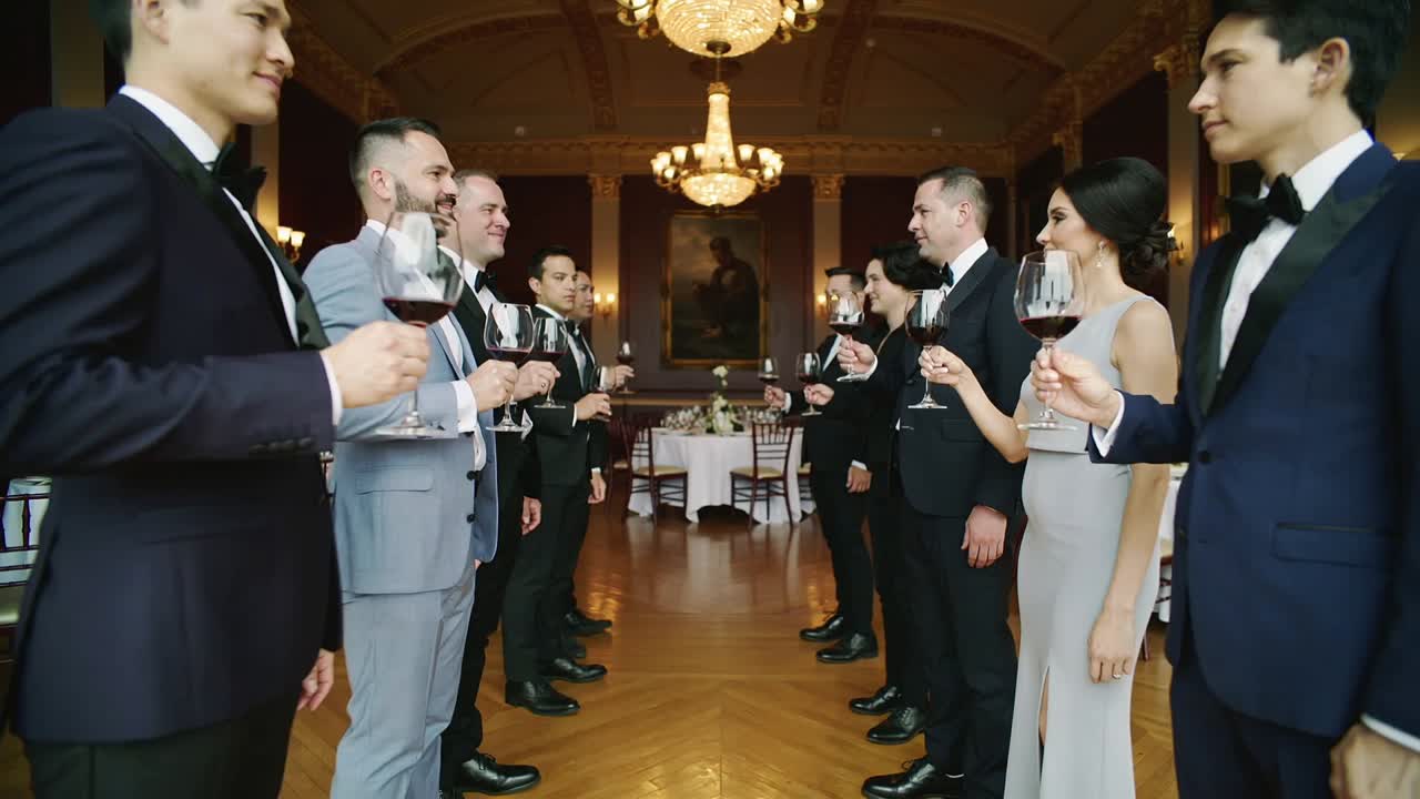 Music cueing guests in two lines, tuxedos, pale gown raising red-wine glasses in hall wedding toast