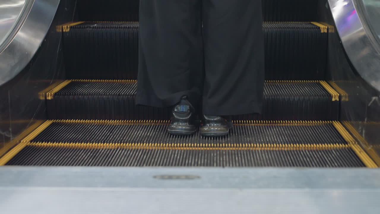 Close up rear view of person wearing black pants stepping onto ascending escalator in indoor public space, showing feet and escalator steps in motion with yellow safety edges and metallic structure