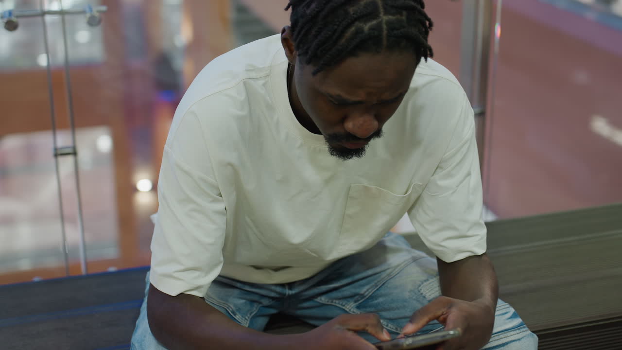 Young man seated in mall chatting over phone with friend dressed in white shirt and jeans looking down at device with blurred escalator light and modern background creating urban lifestyle mood